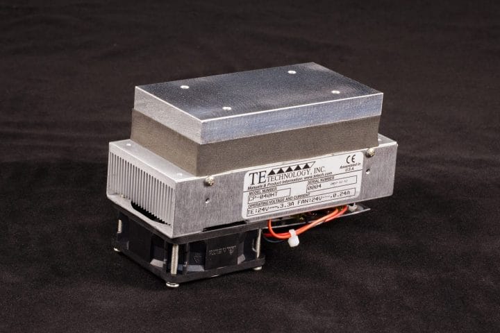 Three-quarter view of 67mm wide high temperature thermoelectric cold plate cooler/heater. Cooled aluminum plate on the front, waste heat exhausted to fins on back. Fins are fan cooled. The fan air enters on the back, off to one side, and exhausts on left and right. Operates on 24 volts DC and removes 40 watts of heat at a 0°C temperature difference relative to ambient. Can heat to 100°C without damaging the cooler. Internal extender blocks improve the capacity at larger temperature differences.